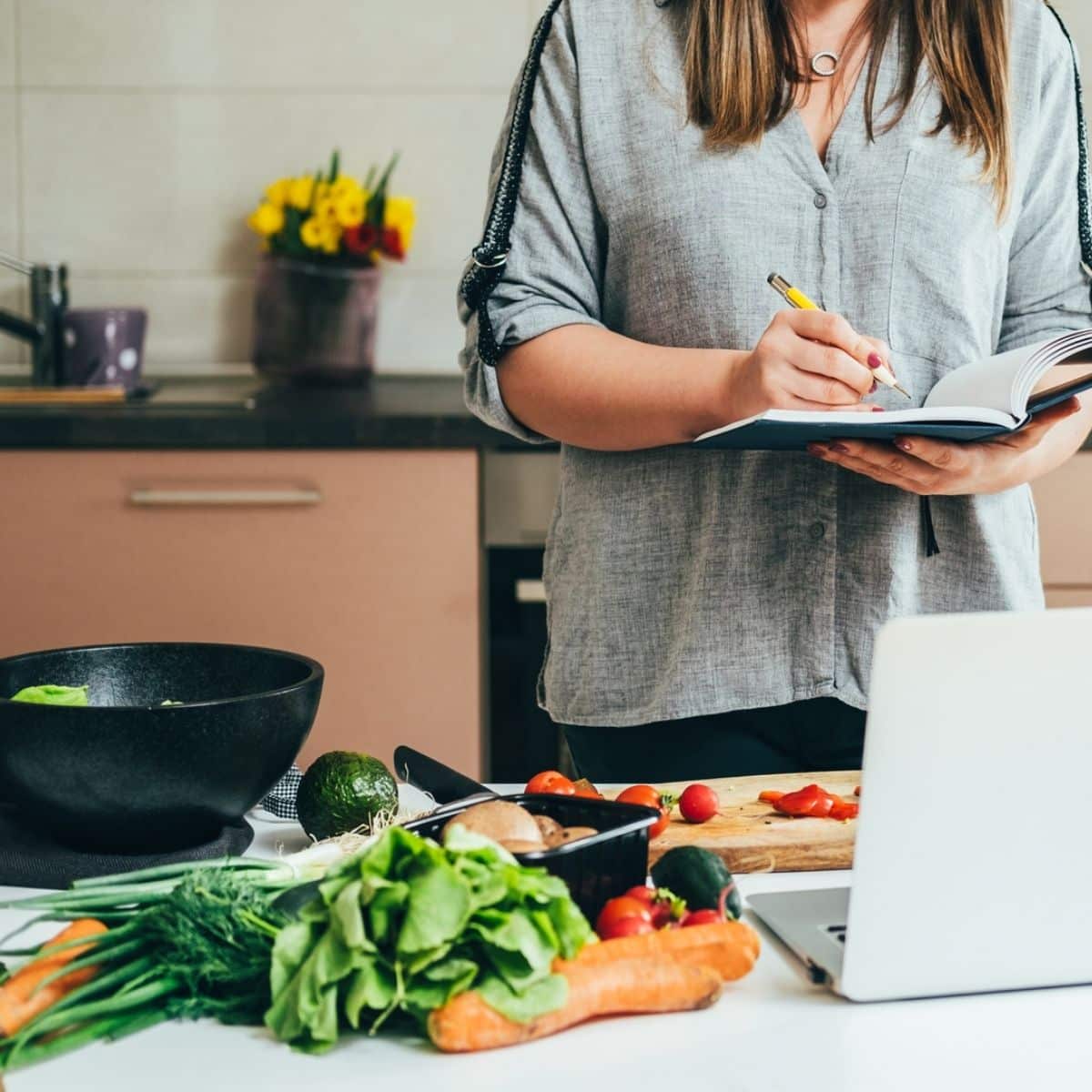 Woman writing new recipe in a notebook while standing at table full with fresh colorful vegetables