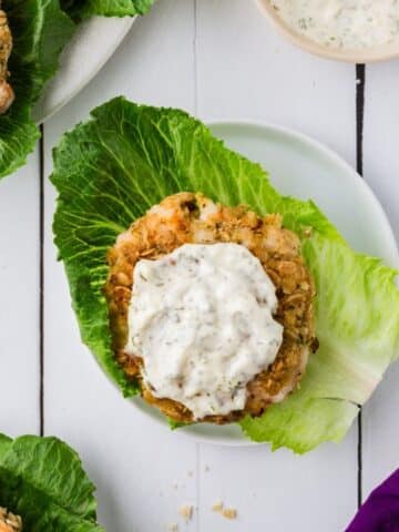 Shrimp burgers on a bed of lettuce with garlic sauce on top. All on a white plate on a white table.