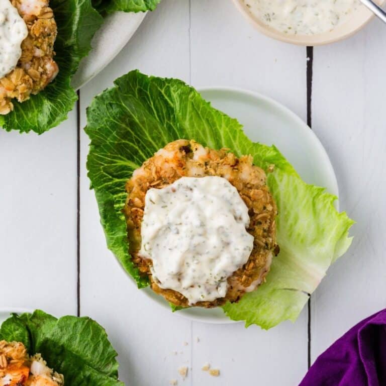 Shrimp burgers on a bed of lettuce with garlic sauce on top. All on a white plate on a white table.